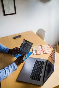 An adult holds a Chinese food menu while using a laptop at a wooden table indoors.