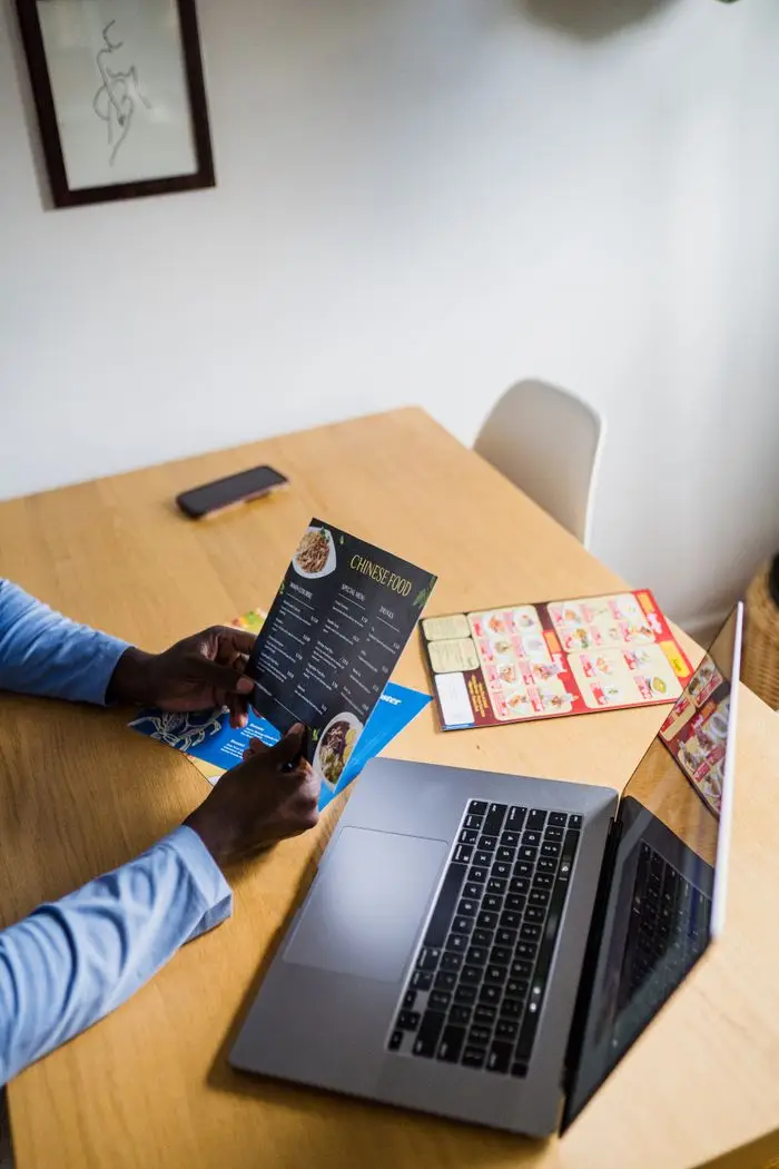 An adult holds a Chinese food menu while using a laptop at a wooden table indoors.