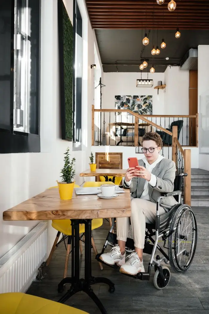 Full body concentrated female in casual wear and eyeglasses using wheelchair surfing mobile phone in modern cafe