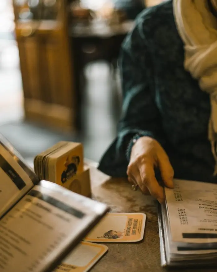Pessoa de blusa escura e cachecol folheando cardápios sobre uma mesa de madeira, em um restaurante aconchegante