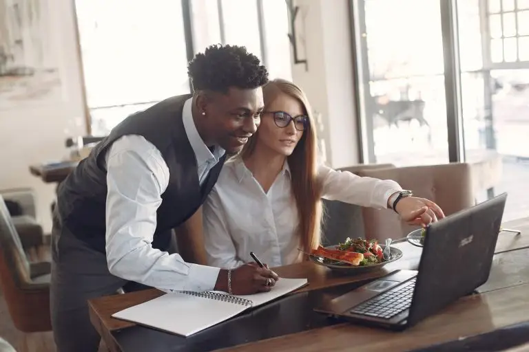 Homem de colete e mulher de óculos olhando juntos a tela de um notebook, com um caderno e um prato de salada sobre a mesa
