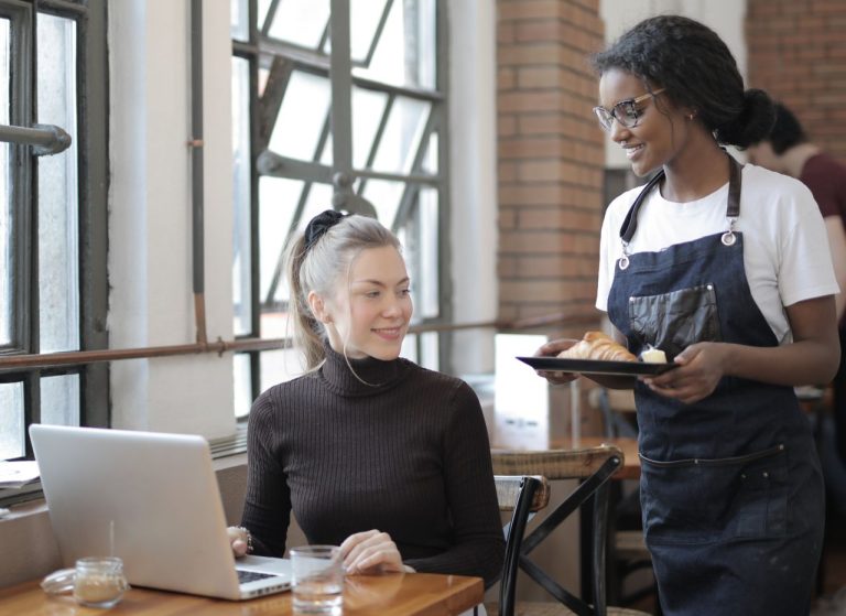 A customer sitting in a café working on a laptop while a waitress serves food, creating a cozy working environment.