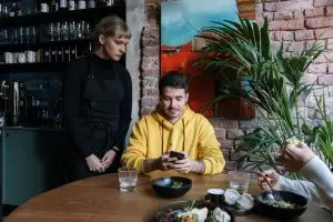 Blog Cliente de blusa amarela usando o celular à mesa, com uma atendente de avental em pé ao lado, em um restaurante