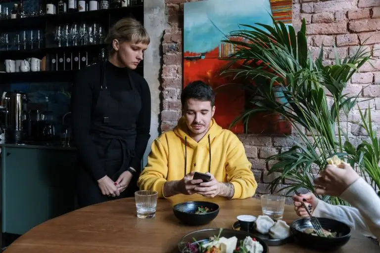 Cliente de blusa amarela usando o celular à mesa, com uma atendente de avental em pé ao lado, em um restaurante